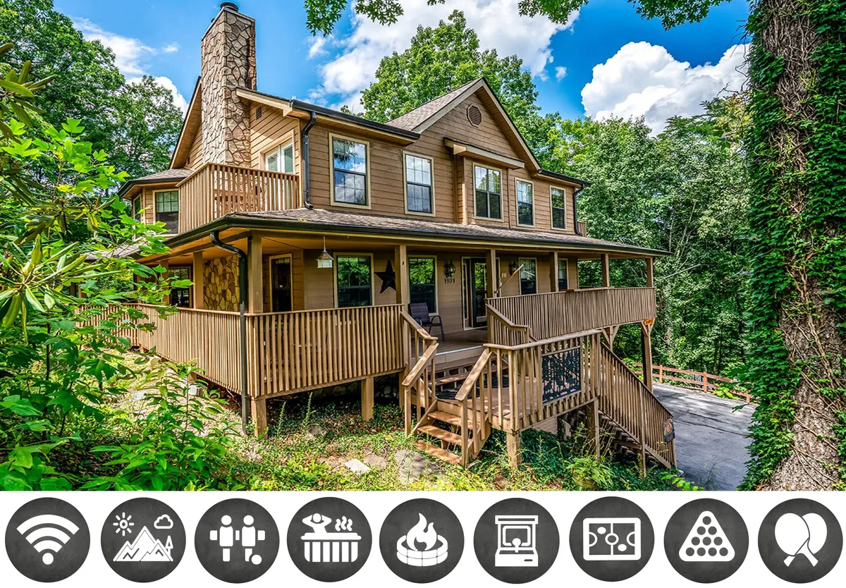 The image showcases the exterior of a beautiful four-story wooden cabin surrounded by lush Smoky Mountain greenery. A spacious deck with comfortable seating wraps around the home, offering views of the sunset. A striking stone chimney hints at a cozy indoor fireplace, while large windows reflect the vibrant natural setting. The inviting porch space is ideal for family gatherings, morning coffee, and relaxing after a day of adventure.