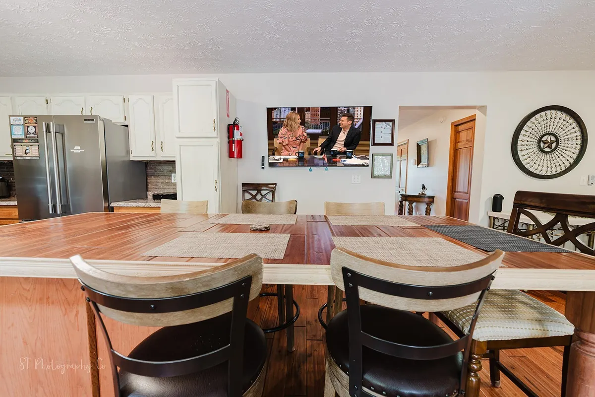 A spacious dining area is centered around a large table, paired with kitchen appliances in the background and a TV with welcoming hosts on display. The scene suggests a space made for lively meals, celebrations, and casual togetherness.
