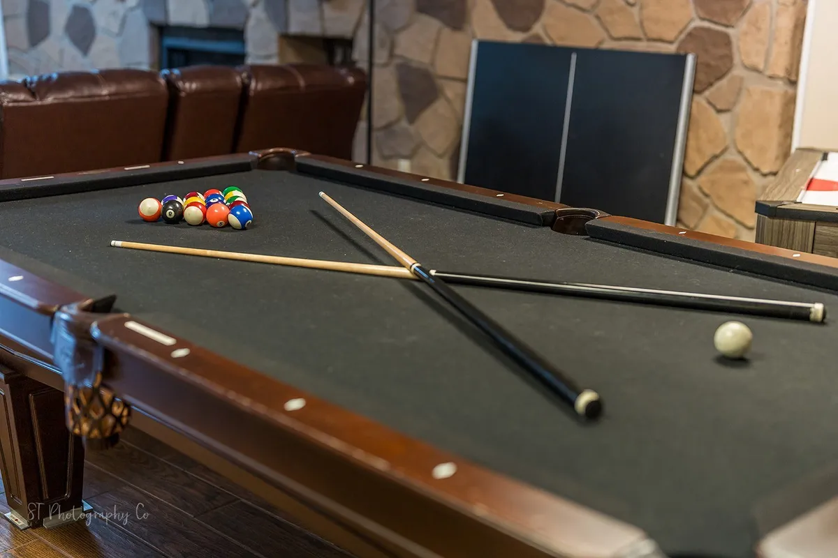 A cozy indoor game room with a pool table, billiard balls, pool cues, a stone accent wall, and a comfortable leather couch for spectators or casual conversations. The casual, friendly atmosphere is perfect for game nights and friendly competitions among groups.