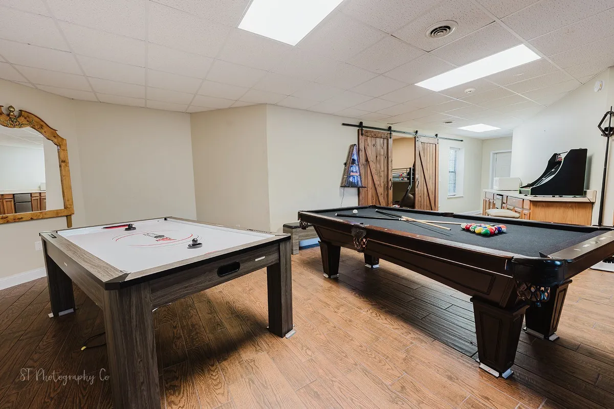 A lively game room featuring a pool table and an air hockey table, both taking center stage in a bright, open space with a large mirror and soft curtains. The layout invites group play and friendly competition, and the atmosphere radiates fun and relaxation.
