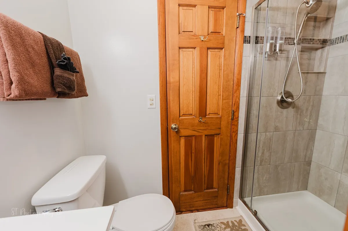 A calming bathroom with a wooden door, glass shower, toilet, and neatly arranged towels. The space feels bright and serene, suggesting opportunities for personal grooming and quiet relaxation.
