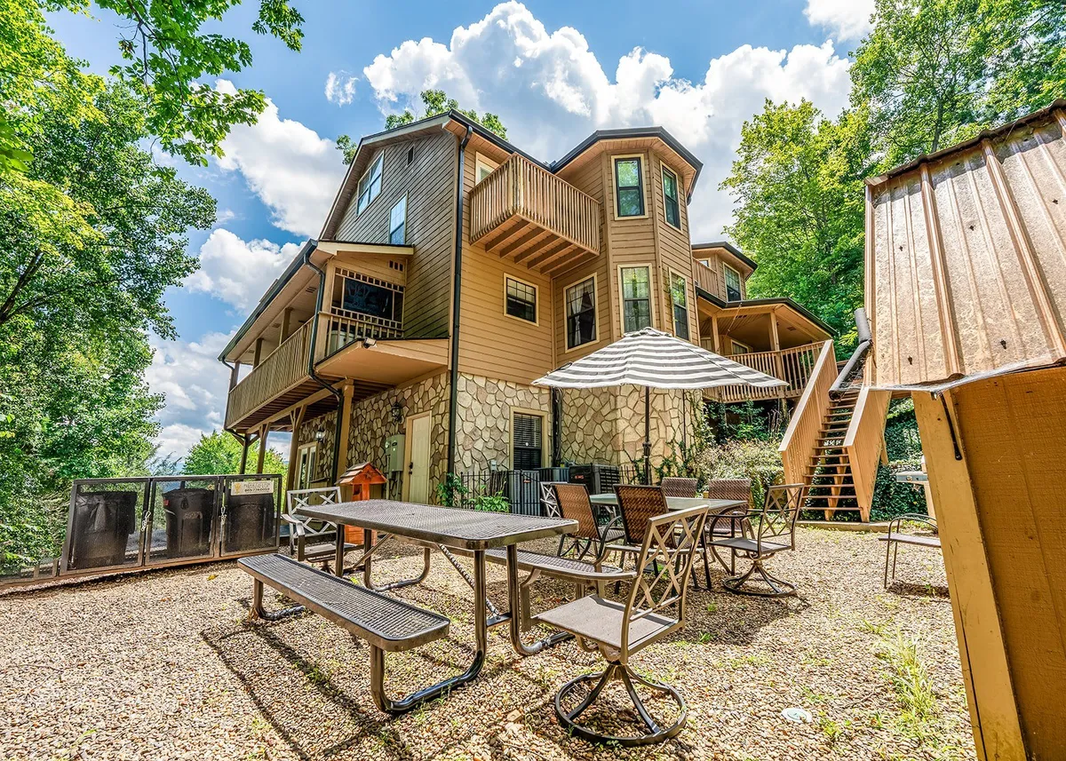 A vibrant exterior view of the multi-level house with an inviting outdoor seating area, an umbrella-covered dining table, and lush trees surrounding the patio. The scene is perfect for communal activities such as outdoor dining, family gatherings, and barbecues, with sunlight filtering through the canopy.
