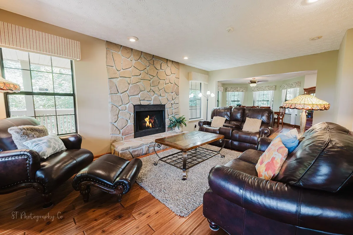This image features a warm, open living room with a prominent fireplace, leather sofas arranged for conversation, a coffee table, and large windows that allow plenty of natural light to fill the space. The inviting layout encourages gatherings and relaxation.
