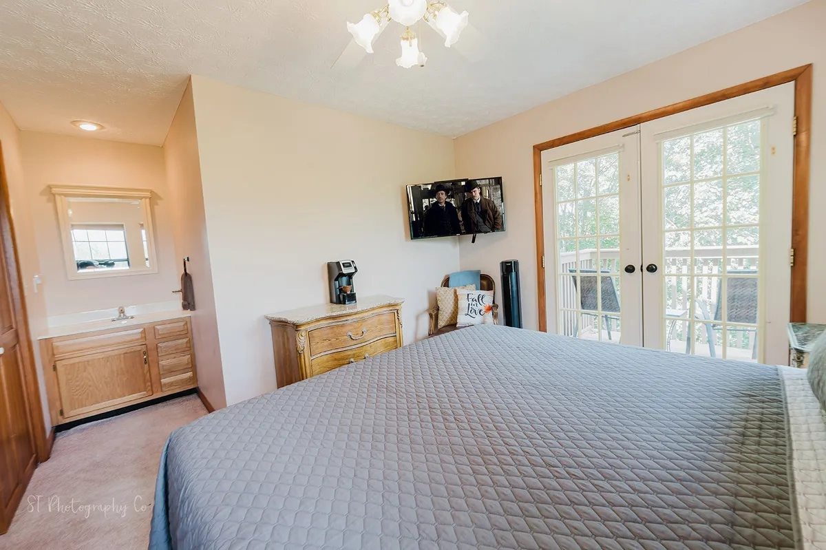 A comfortable bedroom with a bed, TV, vanity, dresser, and, most notably, balcony doors leading outside. The large windows and doors hint at mountain views and easy access to outdoor spaces from the comfort of the bedroom.