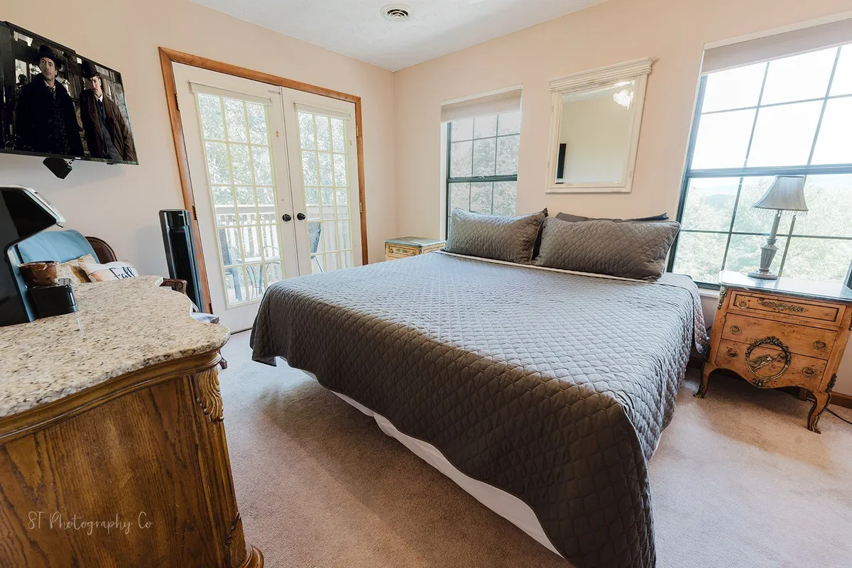 A spacious bedroom featuring a large bed with a gray quilt, two French doors leading to a balcony, a wooden nightstand with a lamp, and a television mounted on the wall. The room is filled with natural light, creating a calm and comfortable atmosphere ideal for relaxation.